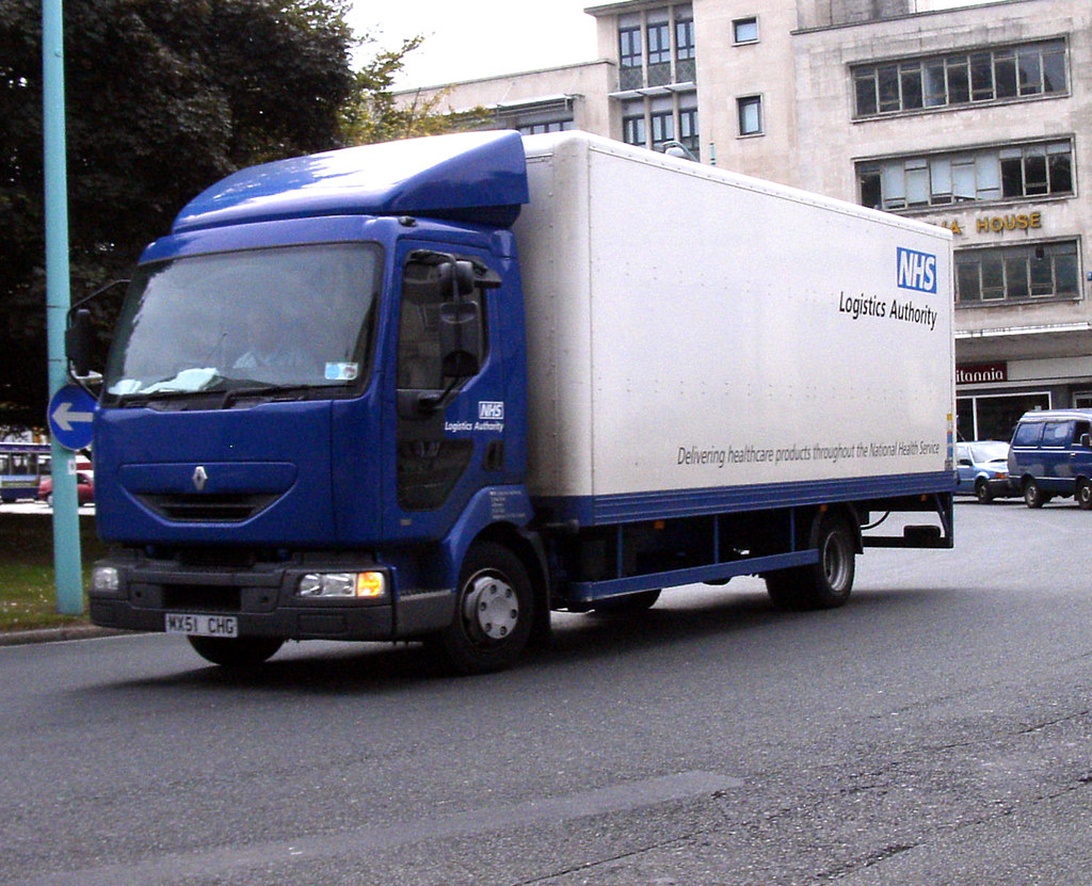 En blå och vit lastbil från NHS Logistics Authority på en stadsgata, med texten 'Delivering healthcare products'.
